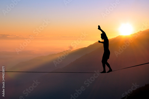 Silhouette of young man balancing on slackline high above clouds and mountains. Slackliner balancing on tightrope during sunset, highline silhouette.
