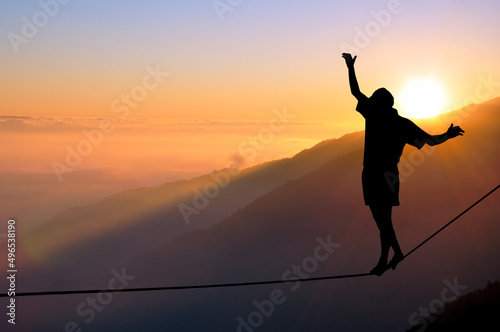 Silhouette of young man balancing on slackline high above clouds and mountains. Slackliner balancing on tightrope during sunset, highline silhouette.