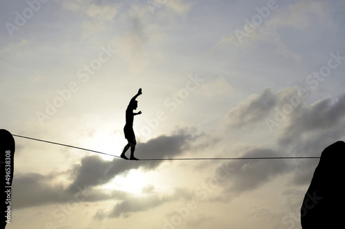 Silhouette of young man balancing on slackline, sun and clouds behind. Slackliner balancing on tightrope between two rocks, highline silhouette.