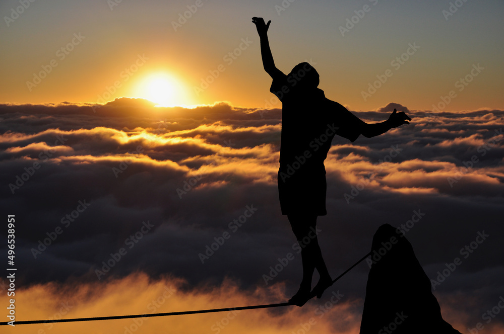 Fototapeta premium Silhouette of young man balancing on slackline high above clouds and mountains. Slackliner balancing on tightrope during sunset, highline silhouette.