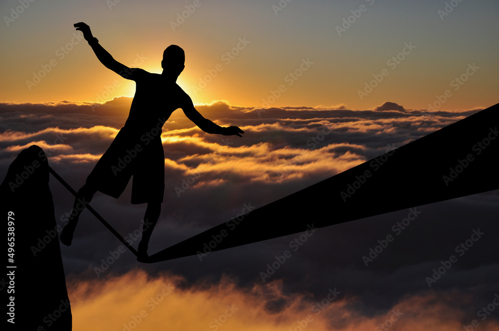 Fototapeta premium Silhouette of young man balancing on slackline high above clouds and mountains. Slackliner balancing on tightrope during sunset, highline silhouette.