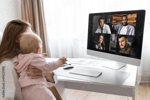 Lovely mother in maternity leave engaged in remote work or study at home on PC while infant baby girl hold documents and looking at screen. Modern day mother of little kid sit at desk use gadgets