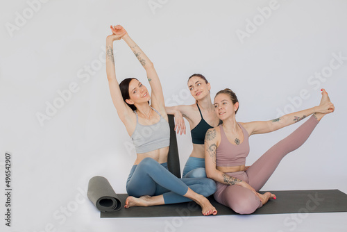 three beautiful girls are relaxing sitting on a yoga mat, reaching out for their hands and talking.
girl sit and stretch her leg in a white studio.
models are dressed in sports leggings and tops