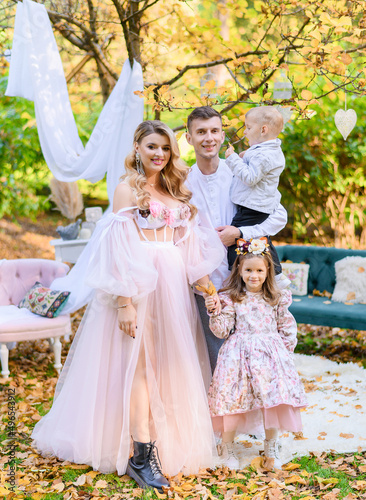 Front view of attractive woman with long curly hair, dressed in pink dress with long sleeves, holding her little daughter by hands, smiling and looking at camera while her husband with son standing © IVASHstudio