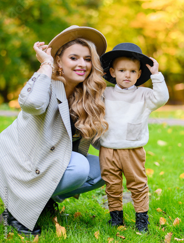 Close view of attractive woman and boy in casual clothes, sitting and holding hats on heads, smiling and posing at camera during sunny autumn day in park