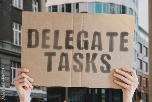 The phrase " Delegate tasks " on a banner in men's hands with blurred background. Office. Develop. Effect. Organize. Organized. Multitask. Help. Teamwork. Successful. Success. Accept. Function. Goal © AndriiKoval