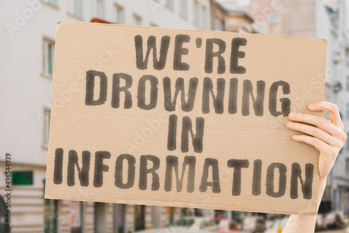 The phrase " We're drowning in information " on a banner in men's hands with blurred background. Stream. Stress. Threat. Abundance. Danger. Spam. Communication. Phone. Device. Answer. Chaos. Confused