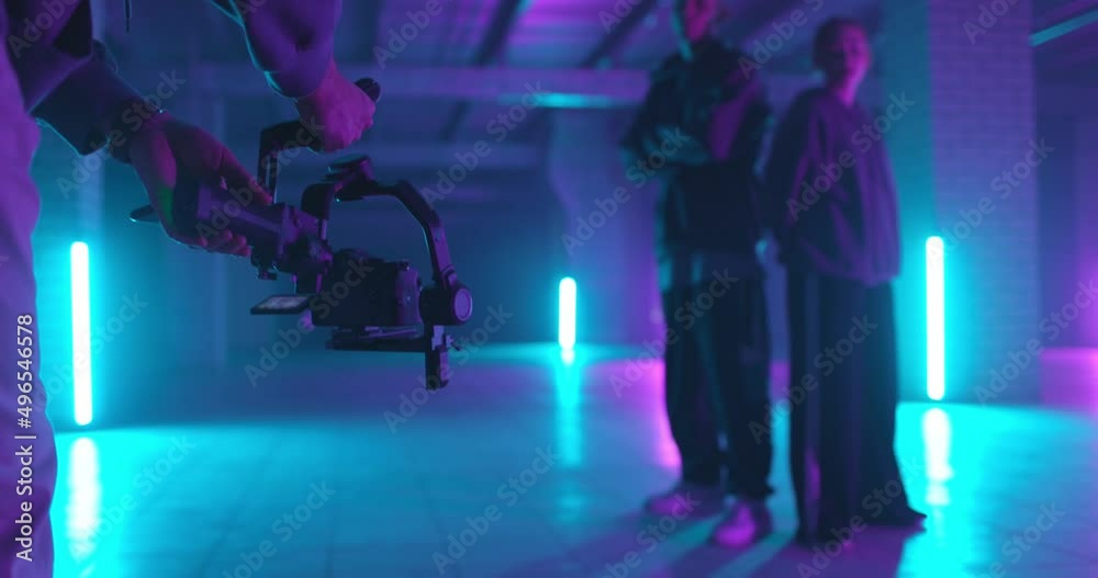 Backstage of video shooting. Cameraman filming hip-hop dancers in neon light. Operator moves with image stabilizer in hands at dark studio. Shooting a clip. Back view