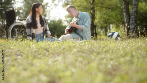 On the background, being in the public park in a good weather, two best friends are sitting on the plaid, a disabled young man is playing guitar and an asian pretty lady with dark long hair is singing
