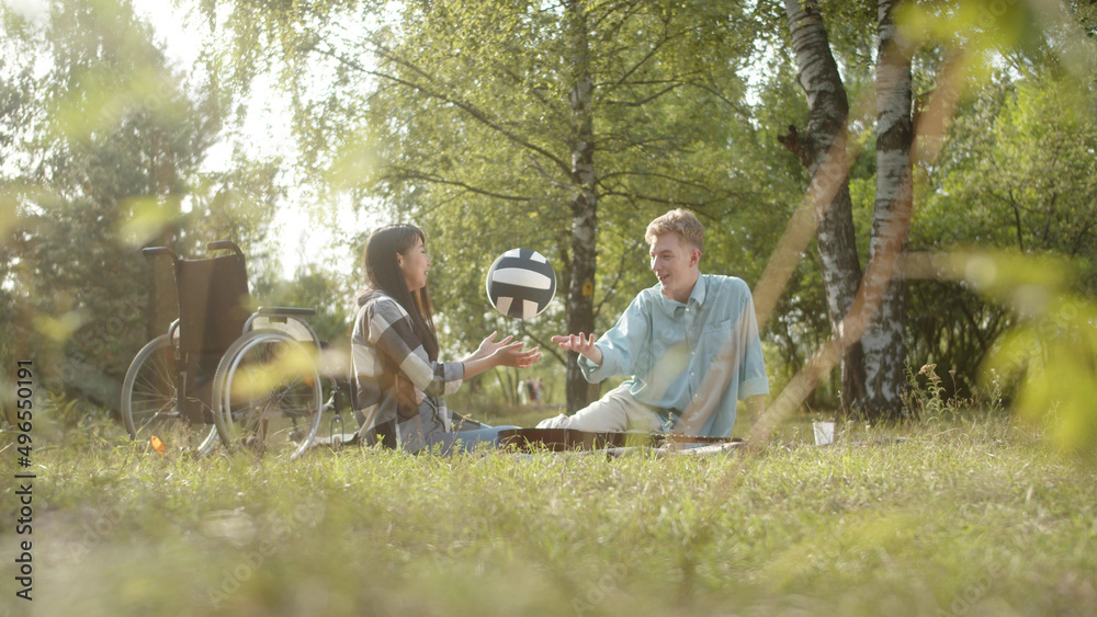 On the background in a good weather two best friends are sitting on the plaid in the public park and throwing the ball to each other On the background in a good weather two best friends are sitting on the plaid in the public park and throwing the ball to each other