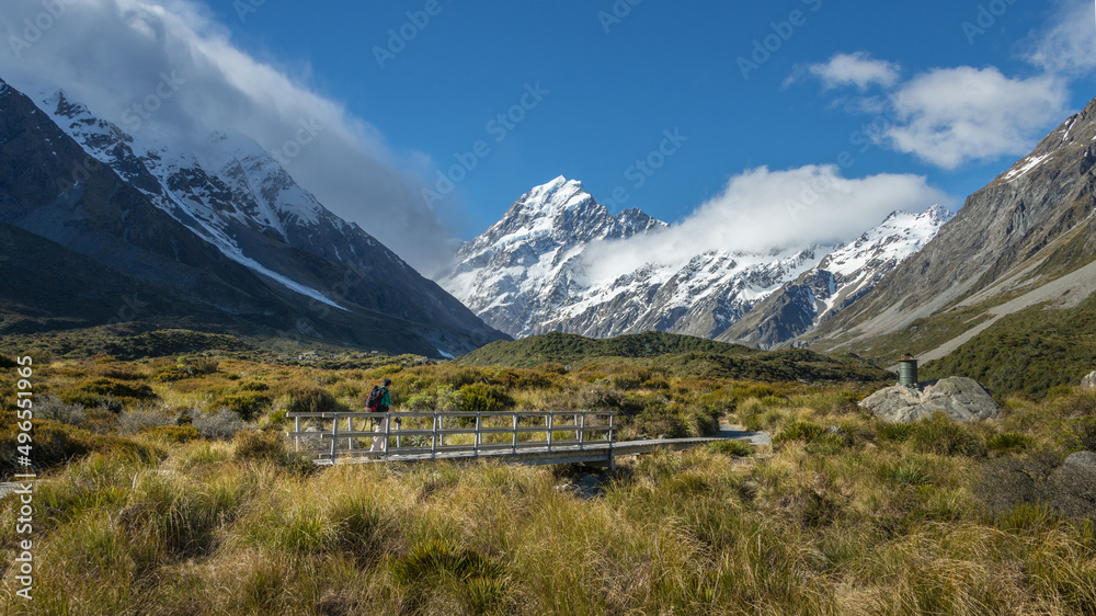 Man walking on a small wooden bridge at Hooker Valley track, Mt Cook in the distance. South Island.