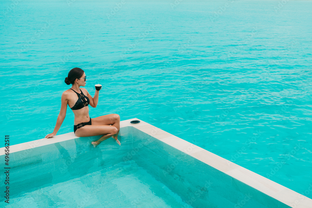 Woman drinking coffee at luxury hotel pool with view of turquoise ocean and overwater infinity swimming pool. Paradise getaway. Fashion model in stylish luxury bikini.