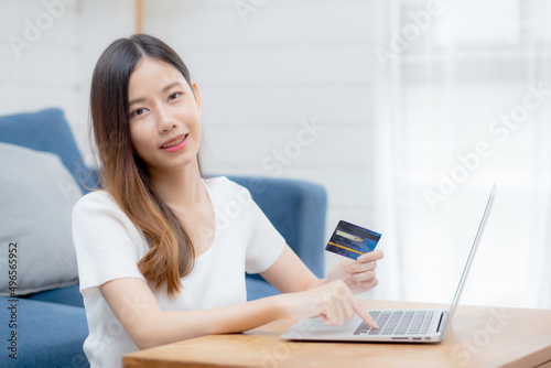 Young asian woman sit on sofa using laptop computer shopping online with credit card buying to internet, happy girl payment with e-business on couch, purchase and payment, business concept. © N_studio