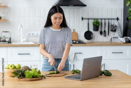 Cooking healthy food concept. Young asian woman watching recipe online using laptop computer while standing at home kitchen photo
