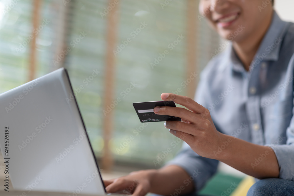 Asian male holding a credit card and smiling at the laptop. Paying for online shopping and services via smart device and computer from home. Young entrepreneur doing things by web communication.