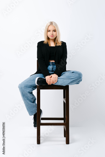 Pretty teenager girl in wide jeans and black jacket sitting on chair with one leg tucked up