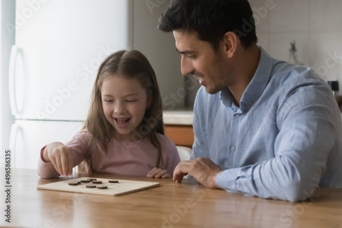 Happy preschool daughter kid and handsome dad playing board game at kitchen table, enjoying home leisure, playtime together. Father teaching cute girl to play checker draughts. Fatherhood concept