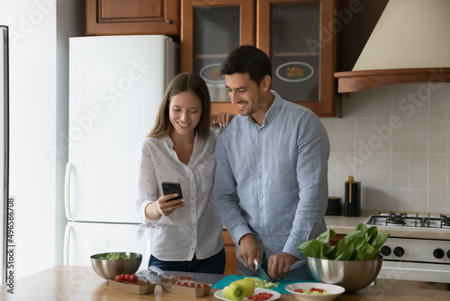 Happy young married couple cooking in home kitchen, checking recipe on mobile phone, ordering food ingredients on internet store. Husband and wife preparing fresh salad for dinner, cutting vegs