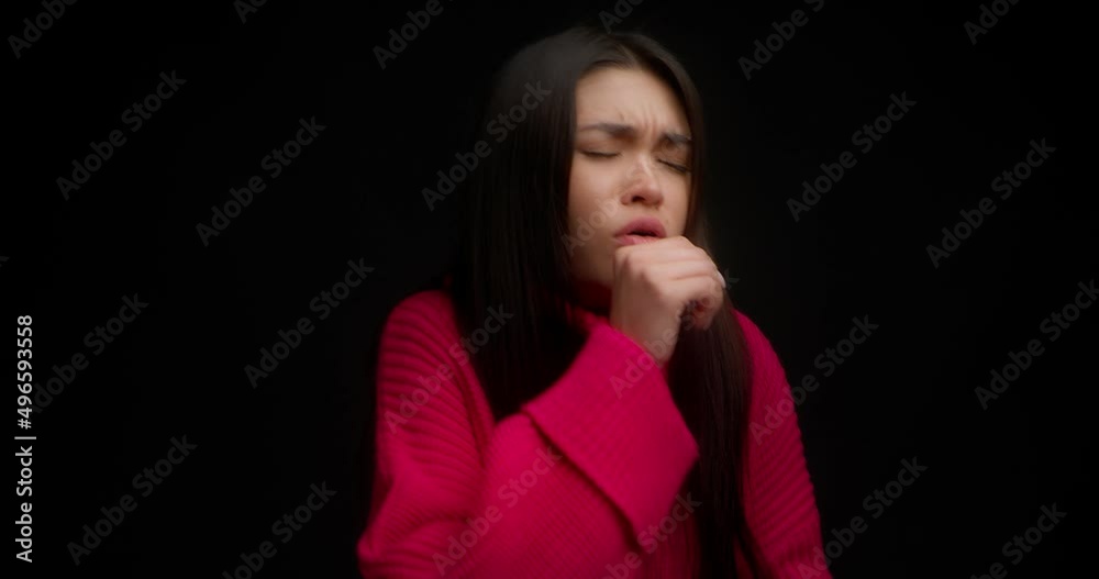 Sick Asian woman in a pink sweater sneezes and coughs covering her mouth with a paper napkin. Runny nose, fever and cough in a young woman isolated on a black background. Sick Asian woman in a pink sweater sneezes and coughs covering her mouth with a paper napkin. Runny nose, fever and cough in a young woman isolated on a black background.