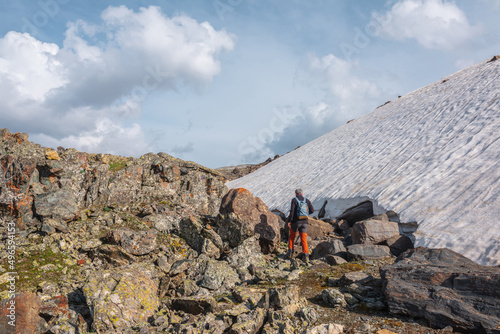 Man walks on moraines along glacier in sunlight under cloudy sky. Tourist in high mountains in sunny day. Traveler on big stones near large glacier in bright sun under clouds in changeable weather.