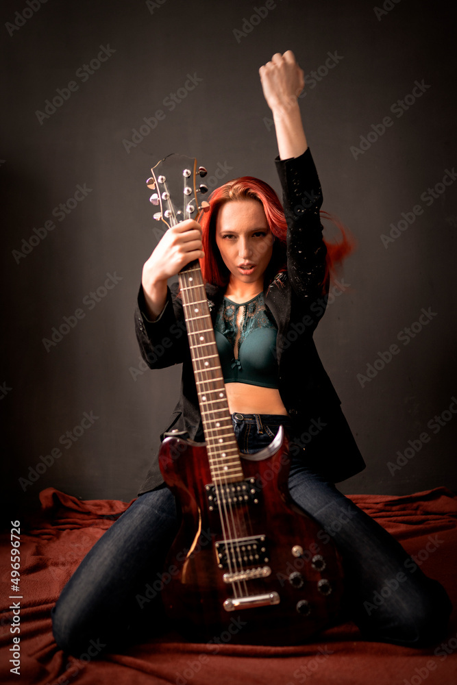 Sexy and brutal rocker punk or metal red-hair girl with an electric guitar in a black jacket and jeans is sitting on the floor on her knees in a defiant pose and hand up in the air