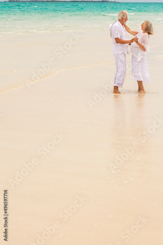Happy mature Caucasian couple in white dancing beside ocean on tropical beach