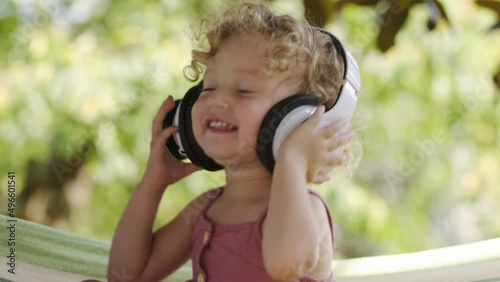 Happy smiling little baby girl listening to music wearing headphones, close-up portrait of funny kid with blue eyes and blond curly hair, laughing child looking at camera, outdoor in sunny summer day.