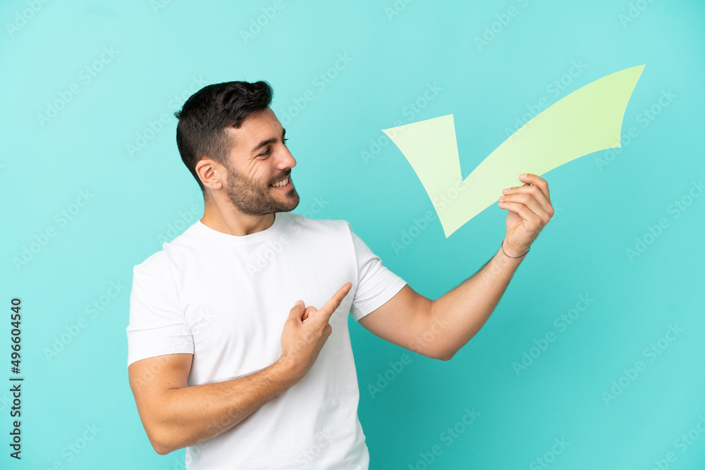 Young handsome caucasian man isolated on blue background holding a check icon and pointing it Young handsome caucasian man isolated on blue background holding a check icon and pointing it