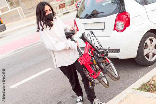 Young teenager with black protective mask has taken his red folding bike out of the car and put it on the sidewalk.