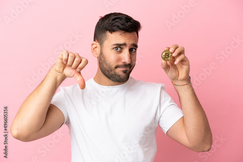 Young man holding a Bitcoin isolated on pink background showing thumb down with negative expression