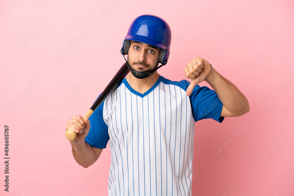 Baseball player with helmet and bat isolated on pink background showing thumb down with negative expression Baseball player with helmet and bat isolated on pink background showing thumb down with negative expression