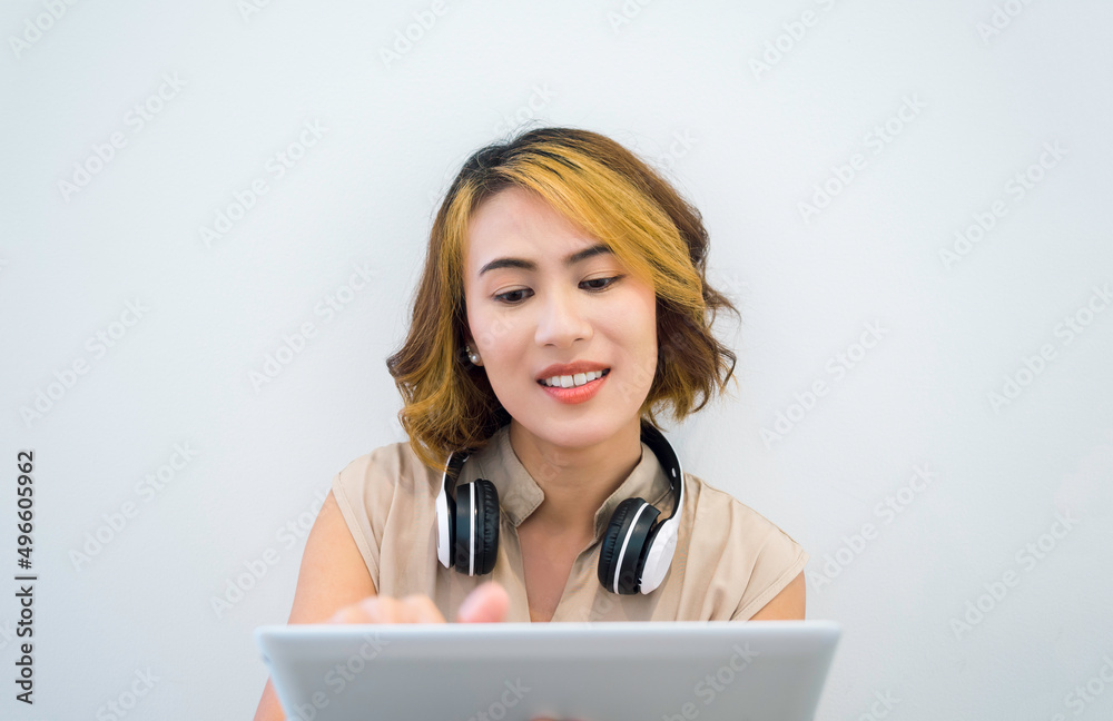 Happy Asian woman short hair in beige sleeveless shirt looking at tablet and touching on screen with white wireless headphone on neck on white wall background. Watching movie, listen music concept. Happy Asian woman short hair in beige sleeveless shirt looking at tablet and touching on screen with white wireless headphone on neck on white wall background. Watching movie, listen music concept.