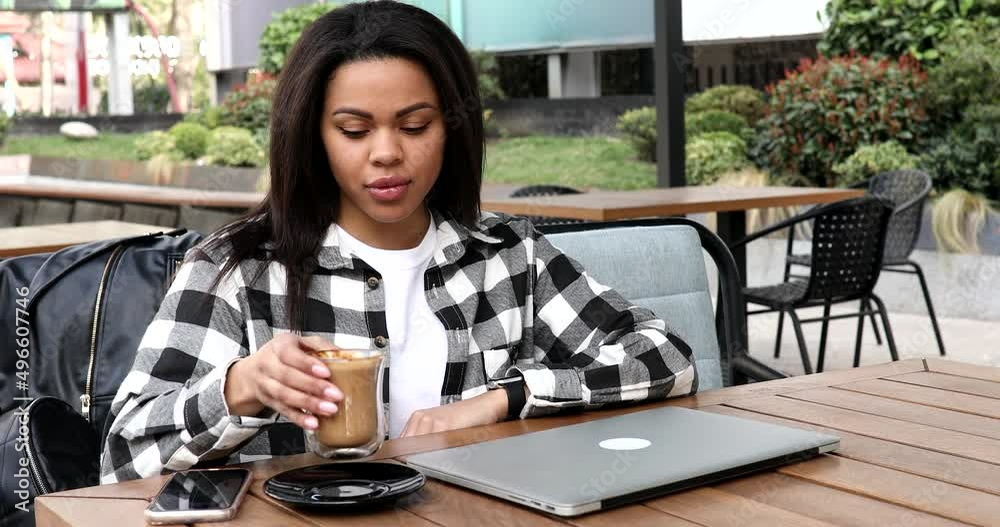Young positive black female in outdoor cafe start working day remotely, opening laptop and typing on it. Young positive black female in outdoor cafe start working day remotely, opening laptop and typing on it.