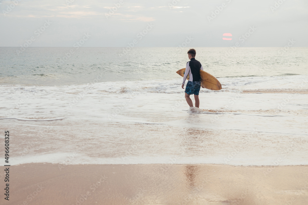 A handsome teenage boy with a surfboard going into the ocean on a sandy beach on the surf line of Sri Lanka.