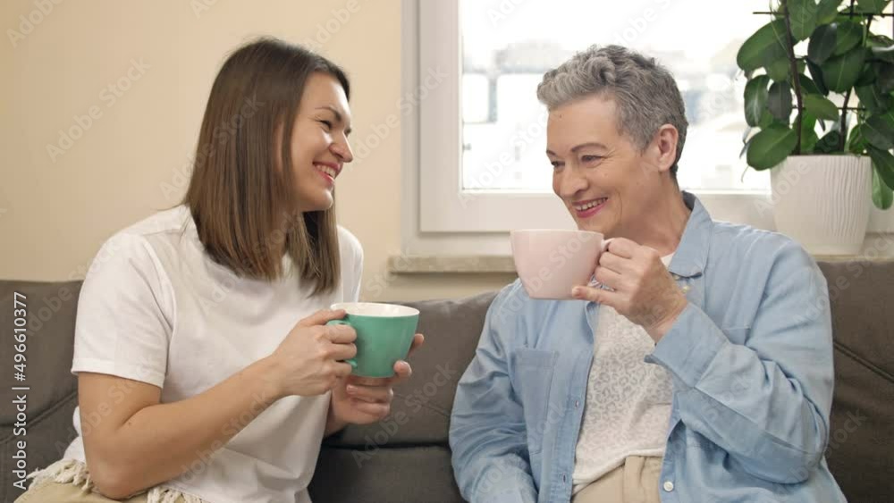 Cheerful women of different ages drink tea or coffee while sitting on the sofa in the living room and have a pleasant conversation. Elderly mother and adult daughter. Mothers Day. Cheerful women of different ages drink tea or coffee while sitting on the sofa in the living room and have a pleasant conversation. Elderly mother and adult daughter. Mothers Day.