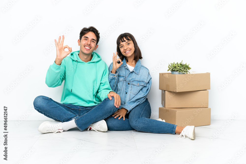 Young couple making a move while picking up a box full of things sitting on the floor isolated on white background showing an ok sign with fingers Young couple making a move while picking up a box full of things sitting on the floor isolated on white background showing an ok sign with fingers