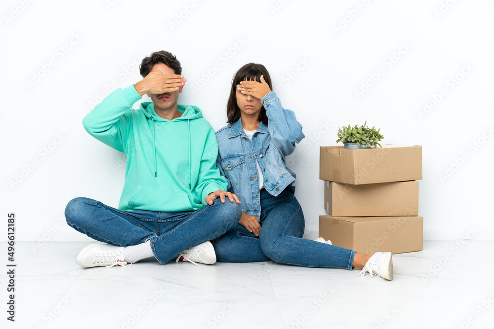 Young couple making a move while picking up a box full of things sitting on the floor isolated on white background covering eyes by hands. Do not want to see something Young couple making a move while picking up a box full of things sitting on the floor isolated on white background covering eyes by hands. Do not want to see something