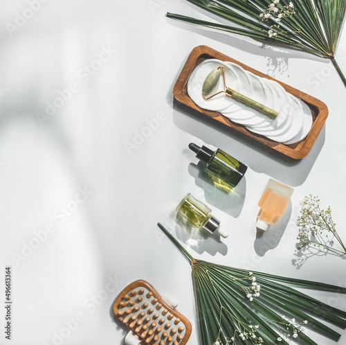 Spa and wellness setting with jade massage roller, massage brush, cosmetic products bottles , flowers and palm leaves on white background with sunlight. Top view.Copy space