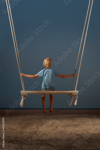 Portrait of boy on swing on the beach on dark sky background. Mystical back view portrait. Concept of childrens loneliness.