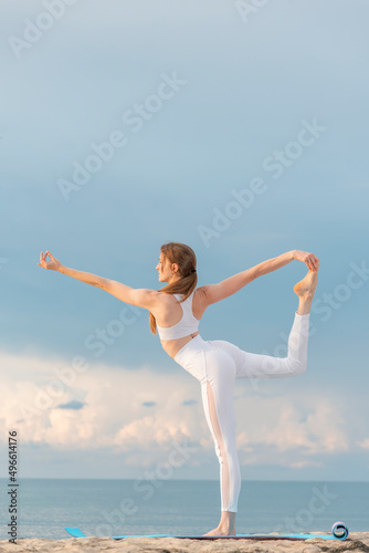 Young woman in white sportwear practicing yoga in the morning on seashore. Doing asana Natarajasana. Yoga at the beach. photo