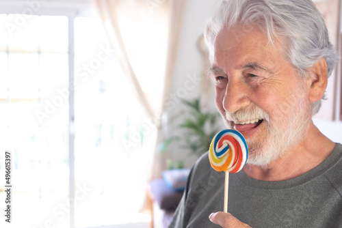 Smiling attractive senior man holding stick with a colorful lollipop with mouth open and tongue out. White-haired grandfather enjoying sweet candies photo