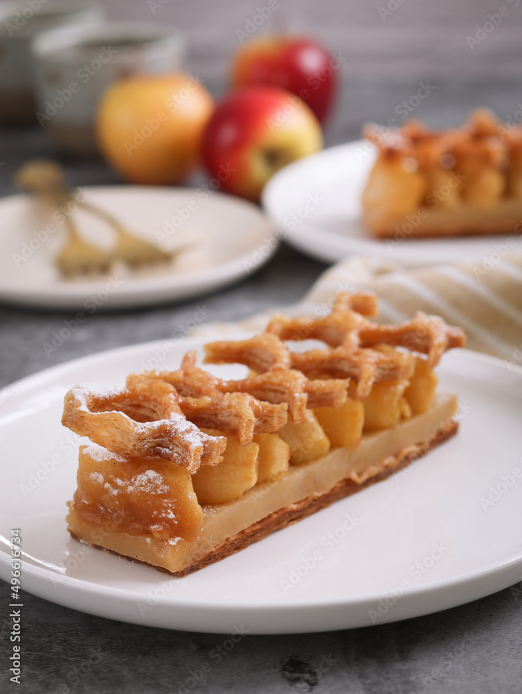 Close up of of traditional caramelize French apple tart, topped with crusty puff pastry pie. With its fruit, plate, forks and cup of tea in background. It's a typical thin tart butter puff pastry Close up of of traditional caramelize French apple tart, topped with crusty puff pastry pie. With its fruit, plate, forks and cup of tea in background. It's a typical thin tart butter puff pastry