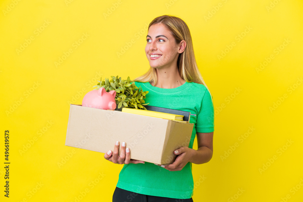 Young Uruguayan girl making a move while picking up a box full of things isolated on yellow background looking to the side and smiling Young Uruguayan girl making a move while picking up a box full of things isolated on yellow background looking to the side and smiling