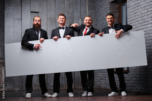 Your advertisement in good hands. Group of handsome young men in toxedo holding big blank with copy space. photo