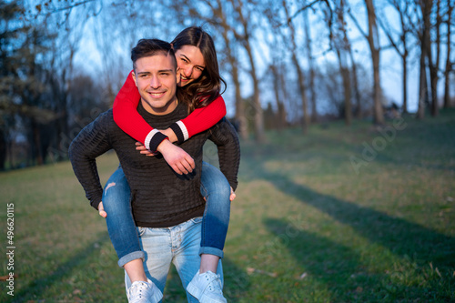 Caucasian couple in spring park smiling, attractive and happy girl hugs handsome athletic guy from shoulders. He moves hloding her piggyback, couple outdoors.