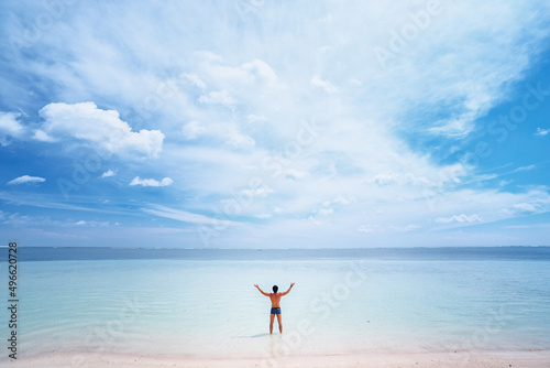 Vacation and freedom. Happy young man rising hands up standing on tropical beach enjoying beautiful view.