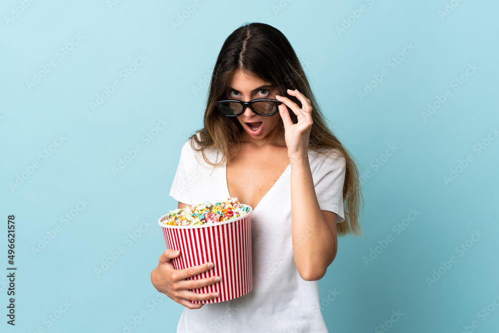 Young caucasian woman isolated on blue background surprised with 3d glasses and holding a big bucket of popcorns