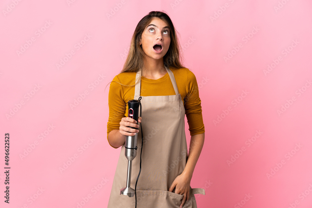 Young chef woman using hand blender isolated on pink background looking up and with surprised expression Young chef woman using hand blender isolated on pink background looking up and with surprised expression