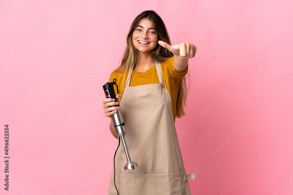 Young chef woman using hand blender isolated on pink background giving a thumbs up gesture Young chef woman using hand blender isolated on pink background giving a thumbs up gesture