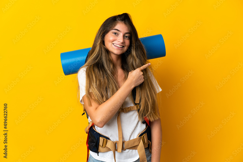 Young mountaineer woman with a big backpack isolated on yellow background pointing to the side to present a product Young mountaineer woman with a big backpack isolated on yellow background pointing to the side to present a product